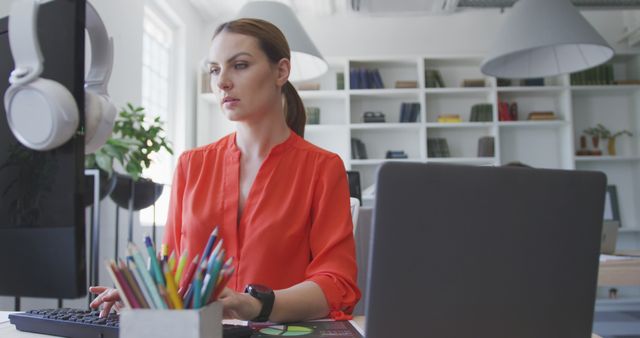 Woman in red shirt working on desktop computer and laptop in modern, bright office. She appears focused and professional. Ideal for business, work-from-home, and productivity themes. May be used in articles about office environments, modern workspaces, or employee engagement.