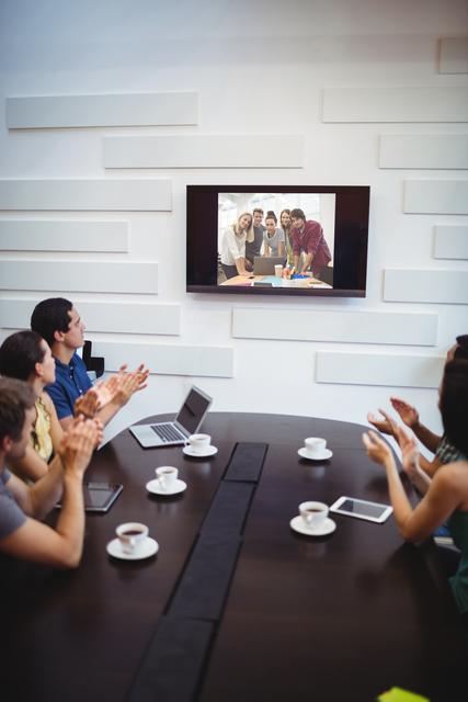 Business executives are seated around a conference table, applauding during a video conference displayed on a wall-mounted screen. This image is perfect for illustrating themes of remote teamwork, modern workplace communication, and corporate meetings. It can be used in business presentations, articles on virtual collaboration, or advertisements for conferencing technology.