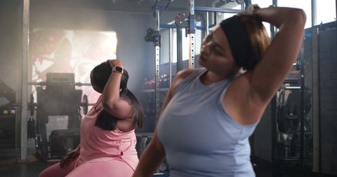 Diverse women doing seated neck stretches in gym near squat rack and weight bench