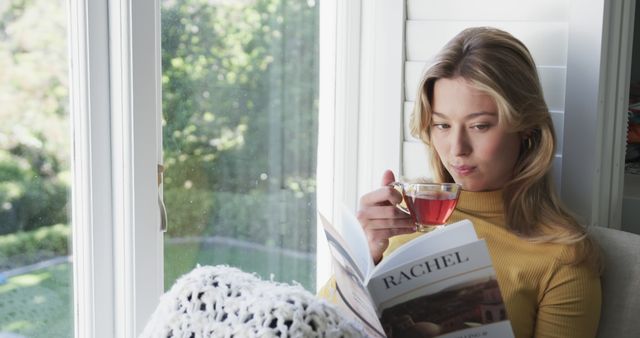Young blonde woman enjoying a relaxing moment by a window in a home setting, sipping tea while reading a book. Suitable for themes of relaxation, home life, leisure activities, and peaceful moments. Great for use in lifestyle blogs, websites promoting wellness and health, or advertisements for relaxation products and services.