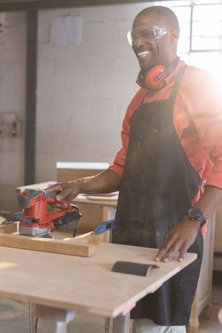 Sanding wooden plank with red electric sander in sunlit carpentry workshop