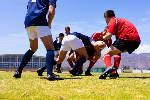 Two teams of multi ethnic male rugby players on a pitch during a rugby game on a sunny day, tackling each other fighting for ball. Sports athletic competition.