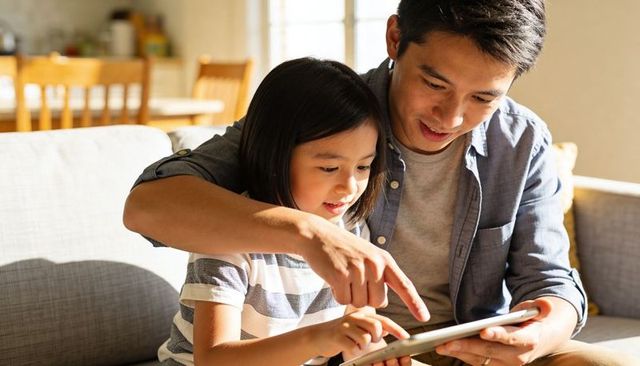 Father guiding daughter tapping tablet on sofa sharing cozy learning moment