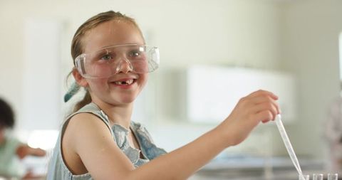 Smiling student wearing safety goggles handling pipette over test tubes in classroom lab