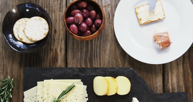 A variety of cheeses are arranged on a slate board accompanied by crackers, olives, and a spread of butter on a wooden table, with copy space. The arrangement suggests a casual yet sophisticated setting for a cheese tasting or a social gathering.