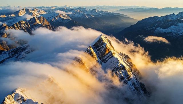 Golden sunrise lighting snow-capped alpine peak rising through misty low clouds