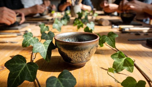 Rustic ceramic teacup sitting on sunlit wooden table with trailing ivy and blurred diners