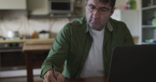 This image depicts a man working from home in a kitchen environment. He is focused on his task, taking notes while using a laptop. This image can be used to illustrate remote work, home office setups, and casual working environments.