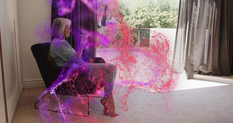 Senior woman sitting in armchair holding cup gazing toward balcony with magenta smoke