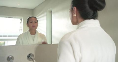 Asian woman in white bathrobe checking skin in bathroom mirror morning skincare routine