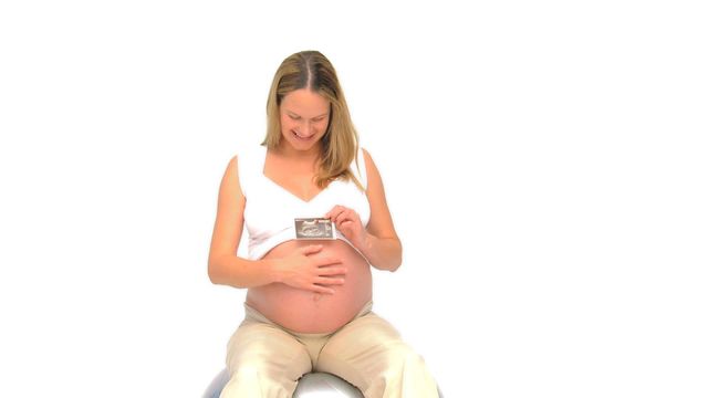 Expectant mother sitting on a gym ball while holding an ultrasound scan, smiling and looking at her belly. Ideal for maternity care, prenatal health, baby products, and family planning advertisements.