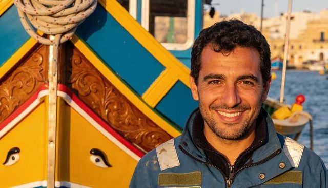 Smiling boatman standing at colorful traditional fishing boat bow with sunlit painted eyes