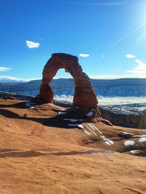 Delicate Arch, located in Arches National Park, Utah, stands magnificently under a bright blue sky with patches of snow around it. Sunlight intensifies the vibrant reddish-orange hue of the sandstone formation. Tourists are visible in the distance, hiking and marveling at the natural wonder. Perfect for travel brochures, nature documentaries, geology studies, hiking gear advertisements, and adventure blogs showcasing scenic American landscapes.