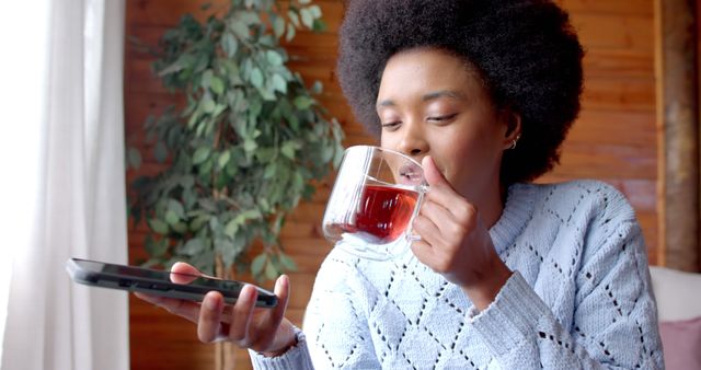 African american woman with afro holding cup of tea and talking on smartphone at home. Lifestyle, free time, communication and domestic life, unaltered.