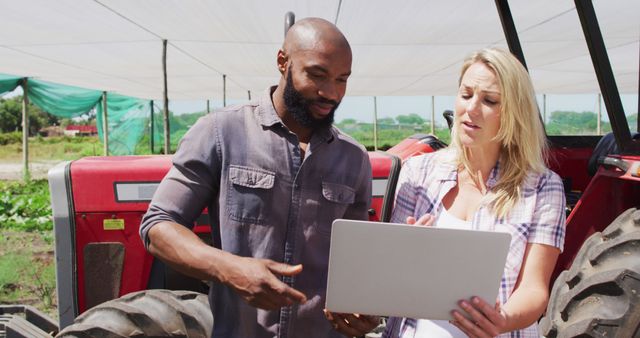 Two farmers discussing farm management using a laptop, standing outdoors near a tractor. Ideal for themes on modern farming, agricultural technology, teamwork, and rural life productivity.