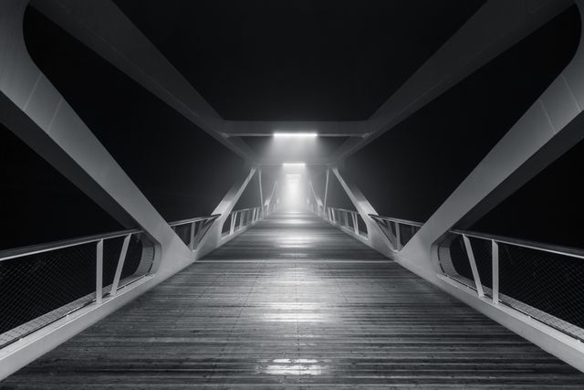 Image showcasing a modern, futuristic pedestrian bridge at night. Dramatic lighting emphasizes the sleek, geometric architectural design. Ideal for concepts related to modern architecture, urban planning, futuristic themes, engineering, and digital wallpapers.