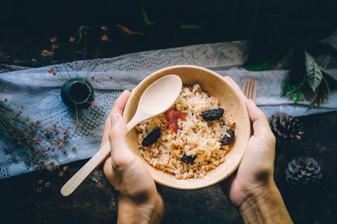 Hands Holding Wooden Bowl of Brown Rice with Dried Fruit, Raisins and Rustic Spoon, Top View
