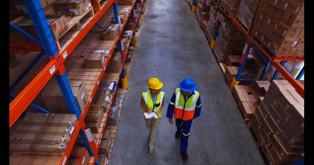 Aerial view of two warehouse workers wearing safety vests and hard hats walking between shelves stacked with packages and boxes. This scene can be used to illustrate concepts of warehouse management, industrial labor, logistics operations and inventory control. Ideal for use in articles about supply chain, safety regulations in industrial environments, and corporate training materials.