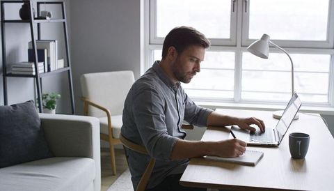 Man writing notes while working on laptop in bright minimalist home office, remote work