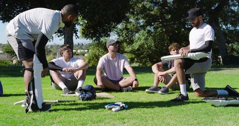 Diverse cricket teammates sitting and kneeling on grass discussing strategy in park