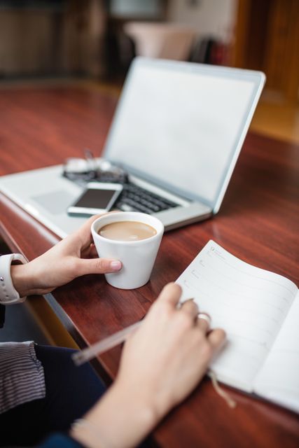 This image shows a woman writing in a notebook while holding a cup of coffee at a desk with a laptop and smartphone. Ideal for use in articles about productivity, studying, remote work, planning, and morning routines. Suitable for educational, business, and lifestyle content.