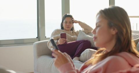 Two Female Friends Lounging on Sofa Using Smartphones in Sunlit Coastal Living Room