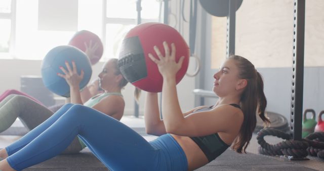 Two women wearing athletic clothing are lying on mats and exercising with medicine balls in a bright and spacious gym. They are performing workouts, focused on strengthening their core muscles. This image can be used for promoting fitness programs, gym memberships, healthy lifestyle blogs, workout training manuals, sportswear advertisements, and physical health campaigns.