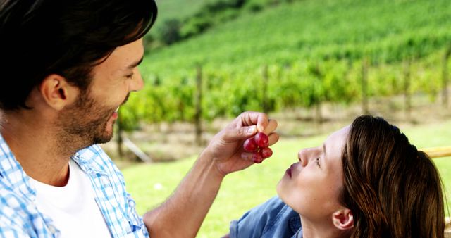 A Caucasian man and woman enjoy a playful moment together in a vineyard, with the man offering grapes to the woman. Their casual attire and joyful interaction suggest a romantic or leisurely day out in the countryside.