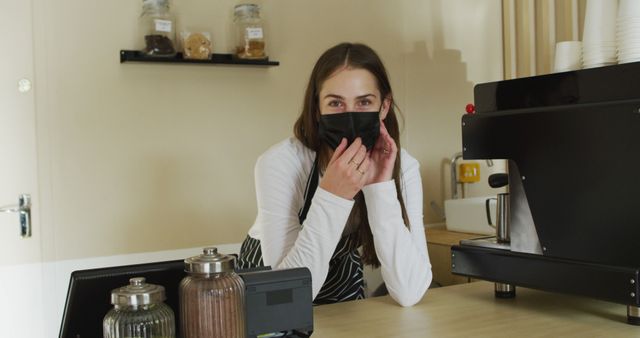 This image shows a friendly barista wearing a face mask standing behind a counter in a coffee shop. She appears welcoming as she tends to her work environment, surrounded by essential coffee-making equipment and jars. Perfect for use in articles or advertisements highlighting safe practices in cafes, small business promotions, and emphasizing hygiene and customer service during pandemics.