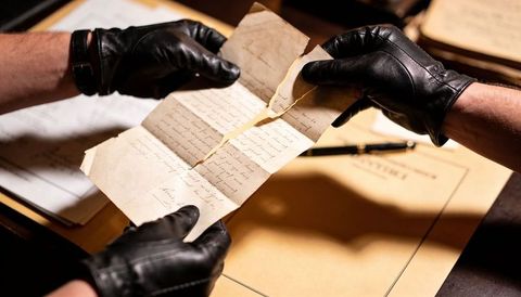 Conservator inspecting torn handwritten manuscript with leather gloves on wooden desk