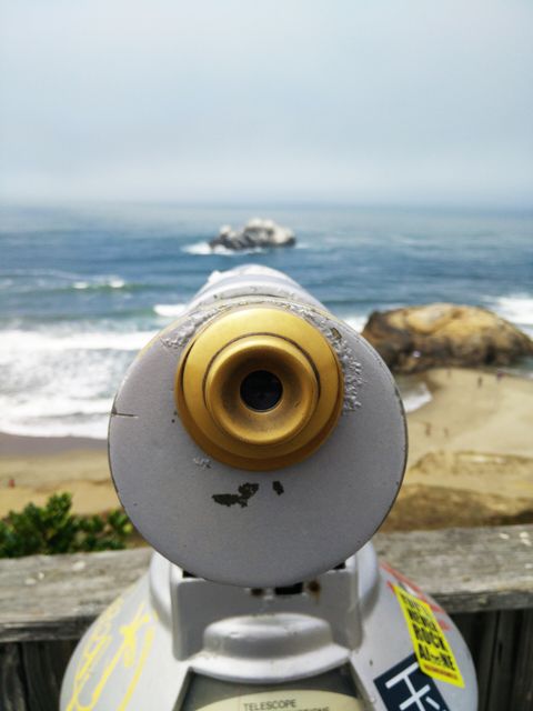 Coin-operated binoculars pointing towards a scenic beach with ocean waves and rocky shore. Useful for travel promotions, tourism ads, website headers for travel blogs or vacation planning, postcards, and scenic promotional materials.