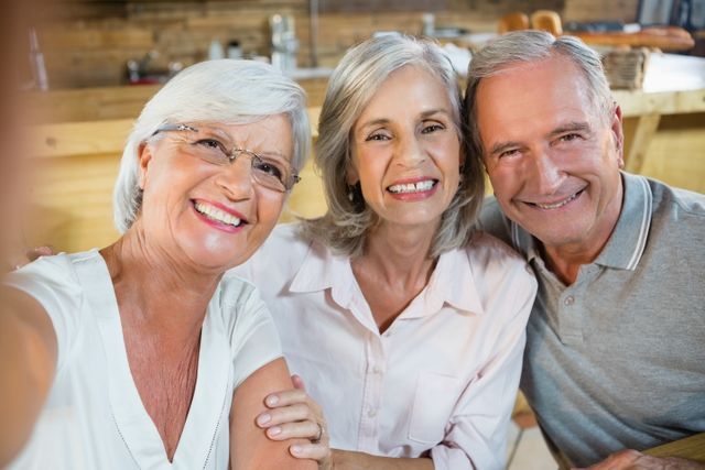 Three senior friends are smiling and posing for a photo in a cozy cafe. They appear happy and relaxed, enjoying each other's company. This image can be used for promoting senior lifestyle, friendship, social gatherings, and retirement activities.
