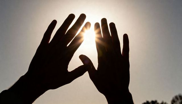Silhouetted hands framing sun with starburst lens flare over clear sky