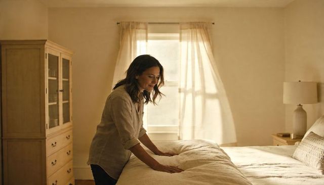 Woman making bed in sunlit cozy bedroom with linen duvet and morning light