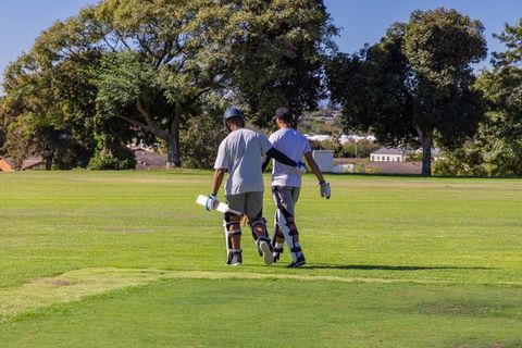 Two cricketers walking across sunlit suburban pitch wearing batting pads and carrying bats