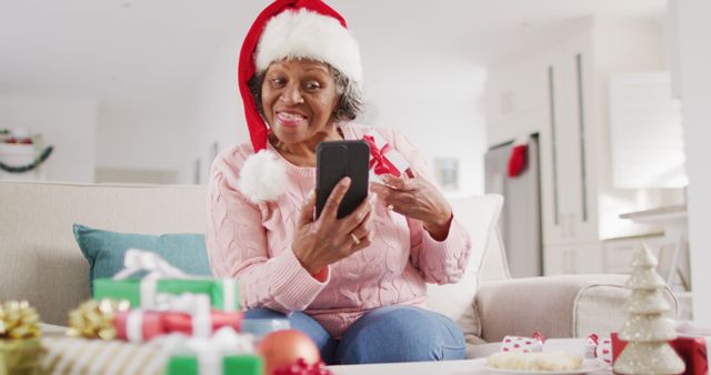 Elderly woman wearing a Santa hat is using a smartphone while surrounded by Christmas gifts. Perfect for holiday celebration themes, elderly and technology, gifts and giving during festive times, and cozy Christmas home settings.