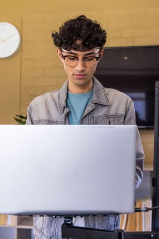 Indian teenage man working at standing desk with laptop and mounted monitor, denim jacket