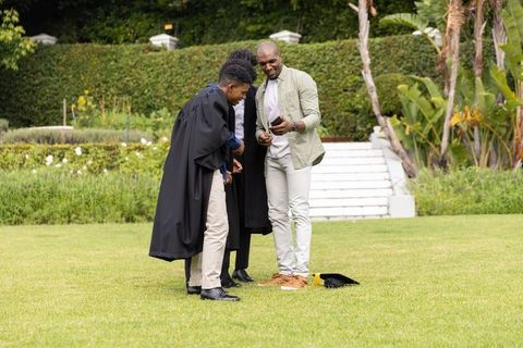 Graduates celebrating on campus lawn viewing smartphone and mortarboard after ceremony