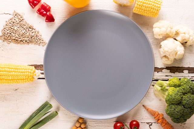 Empty blue plate on white wooden table surrounded by fresh vegetables and seeds. Ideal for healthy eating concepts, organic food promotions, recipe backgrounds, and nutrition blogs.