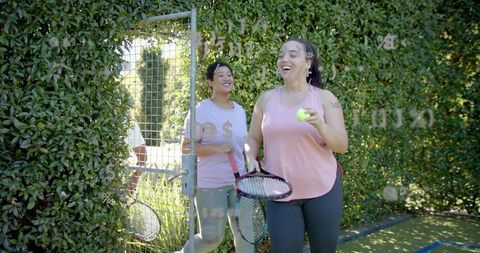 Laughing women leaving hedged tennis court holding rackets and tennis ball, friends