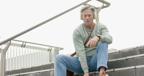 Mature man sitting on urban steps listening with headphones while reaching for shoe