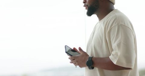 African american man holding smartphone by bright window wearing beanie and watch