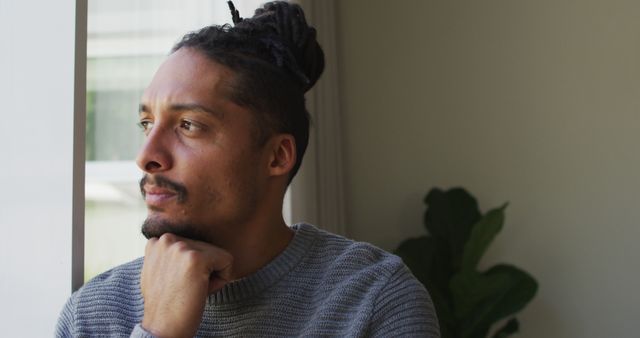 Thoughtful biracial man with dreadlocks in hair bun looking out of window in living room - Download Free Stock Photos Pikwizard.com