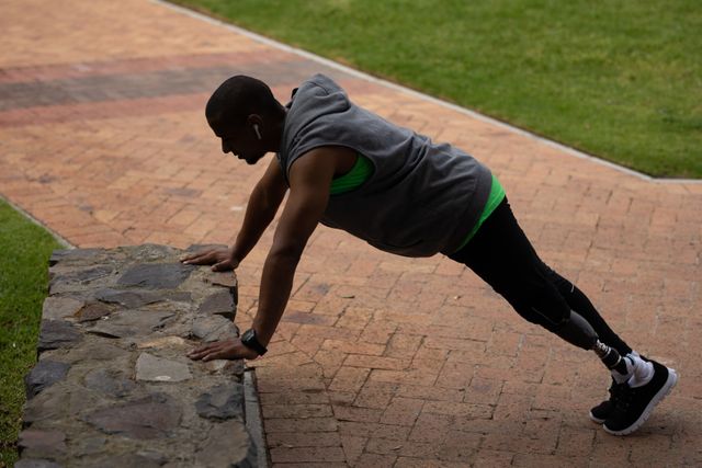 Man with a prosthetic leg exercising in a park, doing push ups while listening to music with wireless earphones. Ideal for content related to fitness, disability, healthy lifestyle, motivation, and outdoor activities. Can be used in articles, blogs, and advertisements promoting inclusive fitness and adaptive sports.