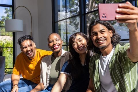 Smiling diverse friends taking selfie on sunlit modern living room sofa, casual hangout