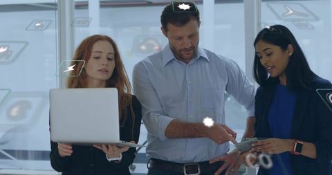 Collaborating team reviewing laptop and tablet in modern glass office with digital icons
