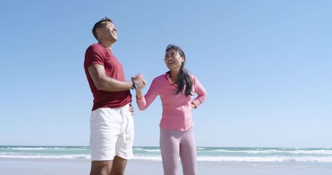 Smiling Diverse Couple Holding Hands on Beach Wearing Activewear and Fitness Trackers