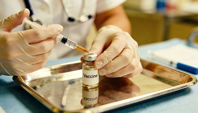 Gloved nurse drawing vaccine into syringe from vial on metal tray preparing immunization
