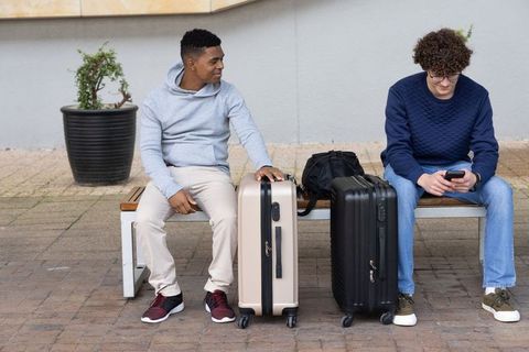 Diverse young friends waiting with rolling luggage on urban bench using smartphone