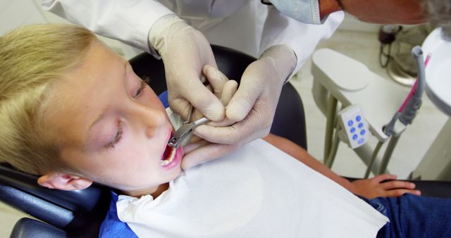 This image depicts a young boy sitting in a dental chair, receiving a dental exam from a dentist wearing white gloves and medical mask. The scene highlights dental care and hygiene, making it useful for articles, websites, and informative content focused on health, pediatric dentistry, or promoting regular dental checkups.